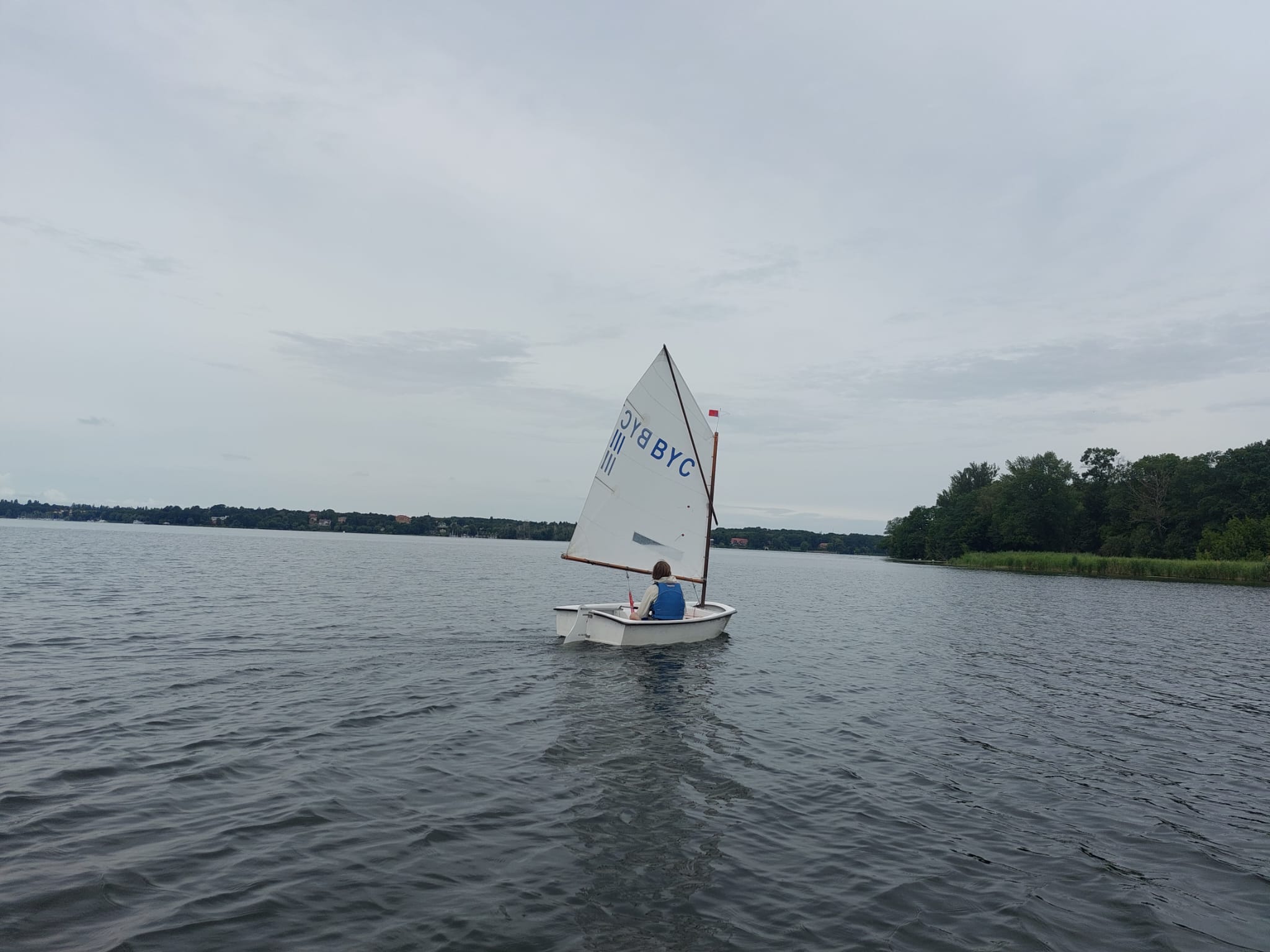 Einzelnes kleines Segelboot mit einer Person auf dem Wasser, im Hintergrund bewaldetes Ufer